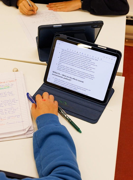 Focus su un iPad nero acceso, affiancato da alcuni fogli scritti a mano e due penne. Al centro, il braccio destro di un bimbo con una felpa azzurra e una macchia rossa sulla mano