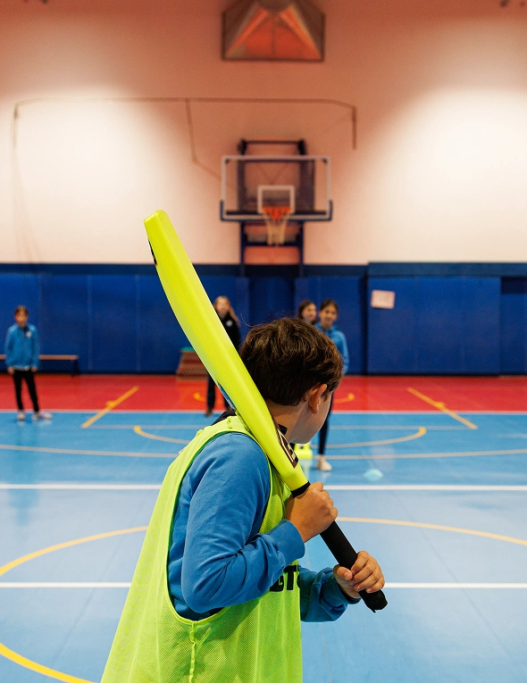 Palestra con pavimento azzurro e campo da basket delimitato da linee gialle e bianche. In primo piano un ragazzo di spalle con un gilet giallo impugna una mazza da baseball gialla con manico nero