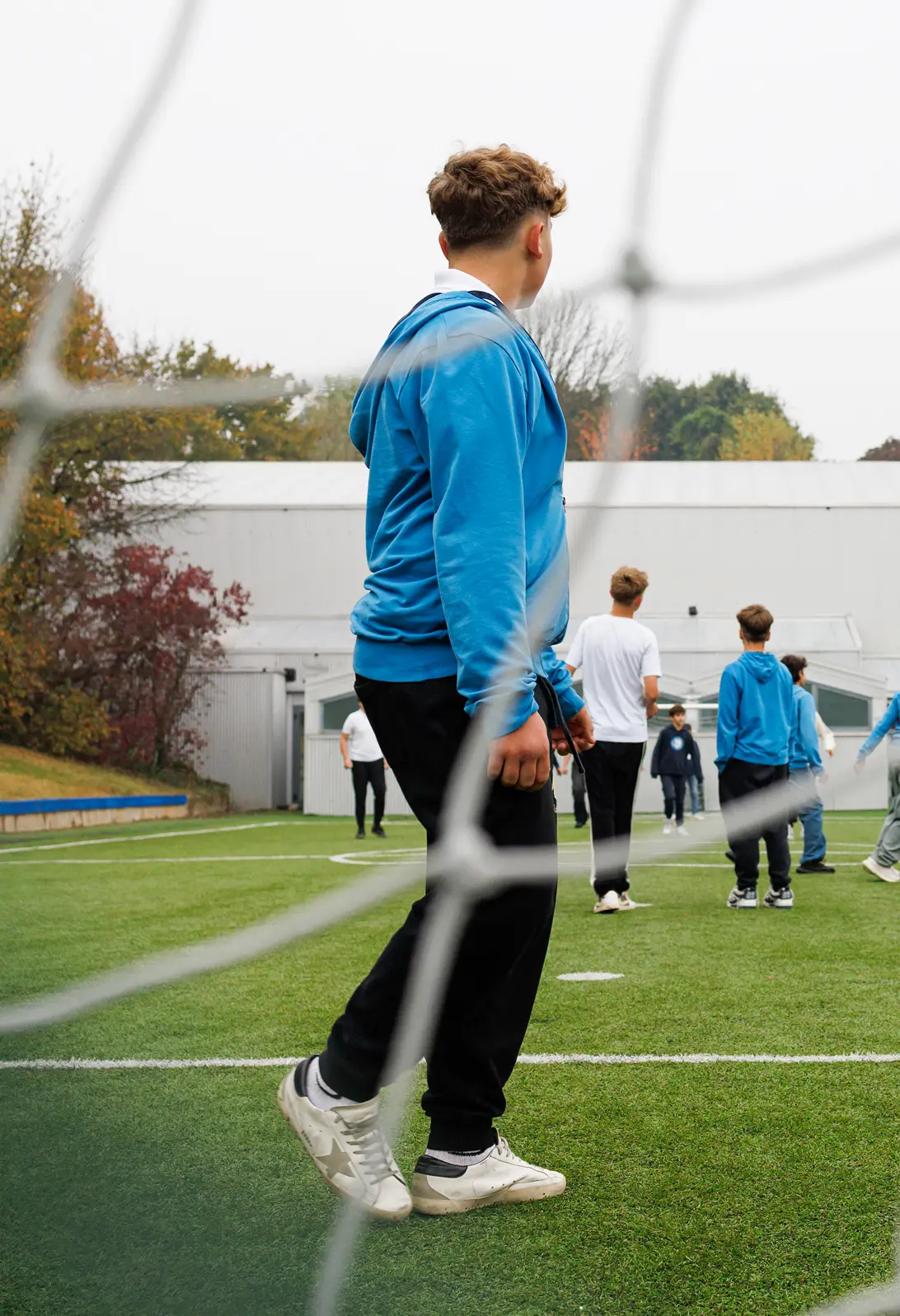 Primo piano di un adolescente con pantaloni neri e felpa azzurra in un campo di calcio, visto attraverso la rete di una porta. Sullo sfondo altri calciatori