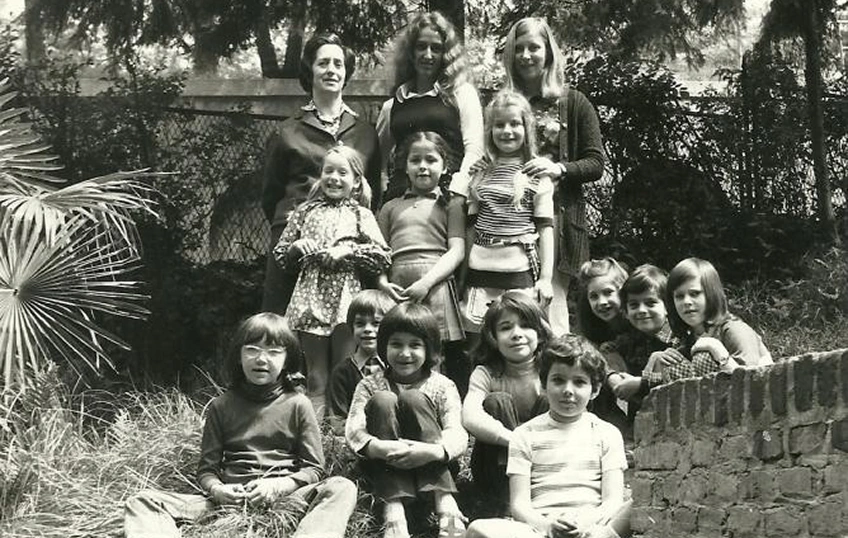 Ritratto in bianco e nero di un gruppo di bambini in età scolare con tre insegnanti nel giardino di IST, scuola internazionale a torino, con palme a sinistra e un muretto in mattoni a destra