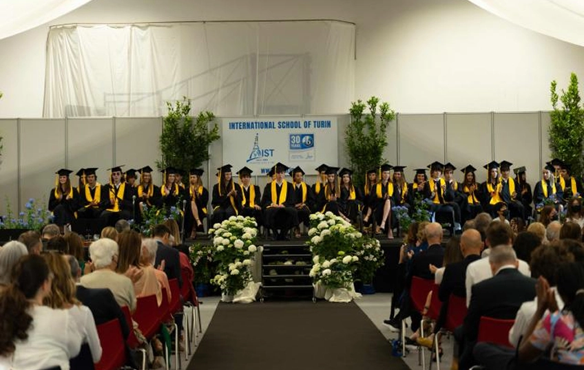 Cerimonia di consegna dei diplomi IB, un numeroso gruppo di diplomandi si trova su una pedana al fondo della sala, con toga e tocco neri e sciarpa gialla. Nella platea due ali di persone sedute applaudono