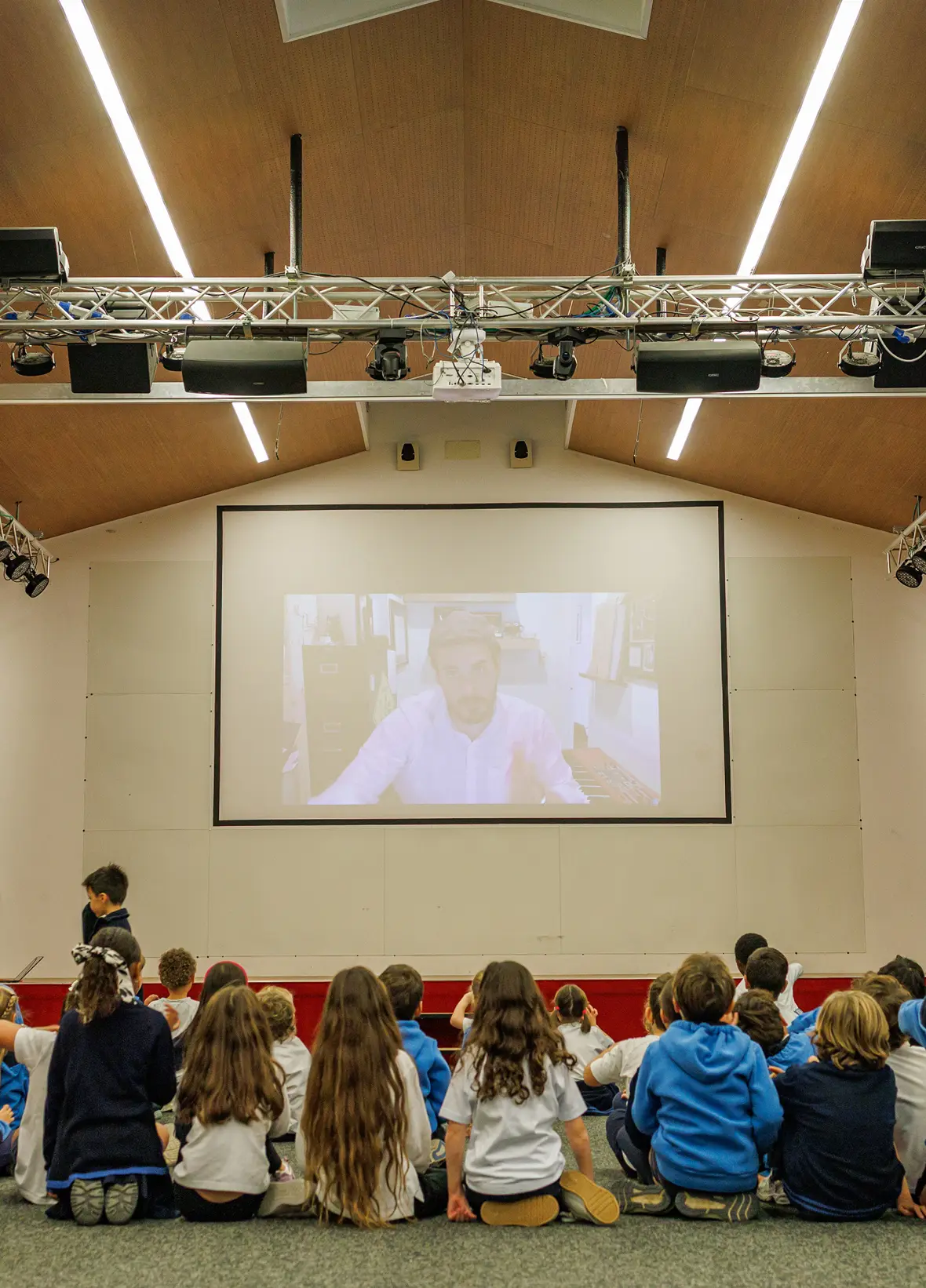 20 bambini seduti a terra in un'aula di proiezione con struttura metallica orizzontale a soffitto assistono a un video sull'international baccalaureate