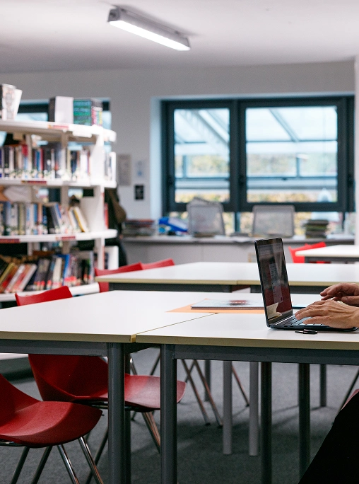 Interno della biblioteca scolastica IST, tavoli beige con gambe grigie, sedie rosse, a sinistra scaffali di libri, sulla destra le mani di una persona che lavora su un computer portatile