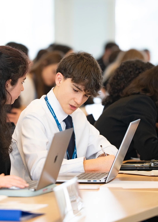 Uno studente utilizza i servizi di supporto scolastico forniti dall’IST, impiegando un computer portatile mentre scrive con una penna. Al suo fianco una ragazza con il laptop aperto