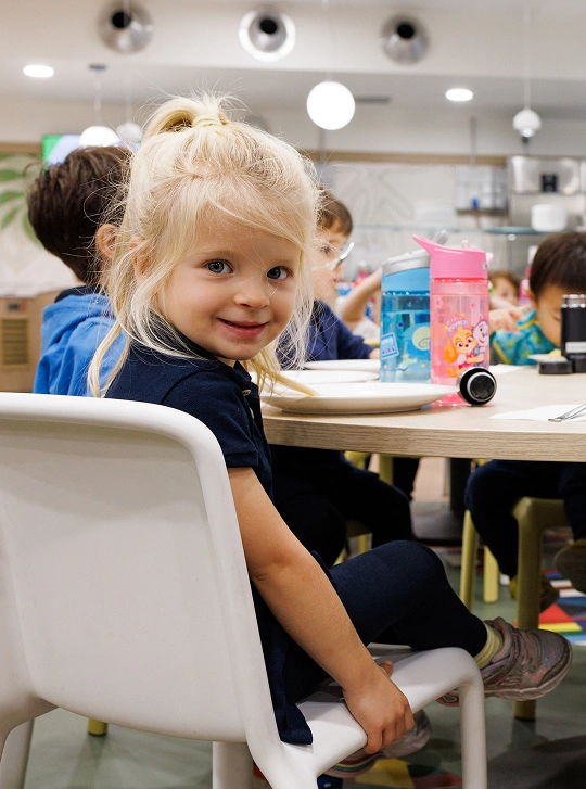 Gruppo di bambini nell’atto di mangiare a una tavola imbandita. La piccola sorridente in primo piano, rivolta verso l’osservatore, ha i capelli biondi raccolti sul capo e gli occhi azzurri