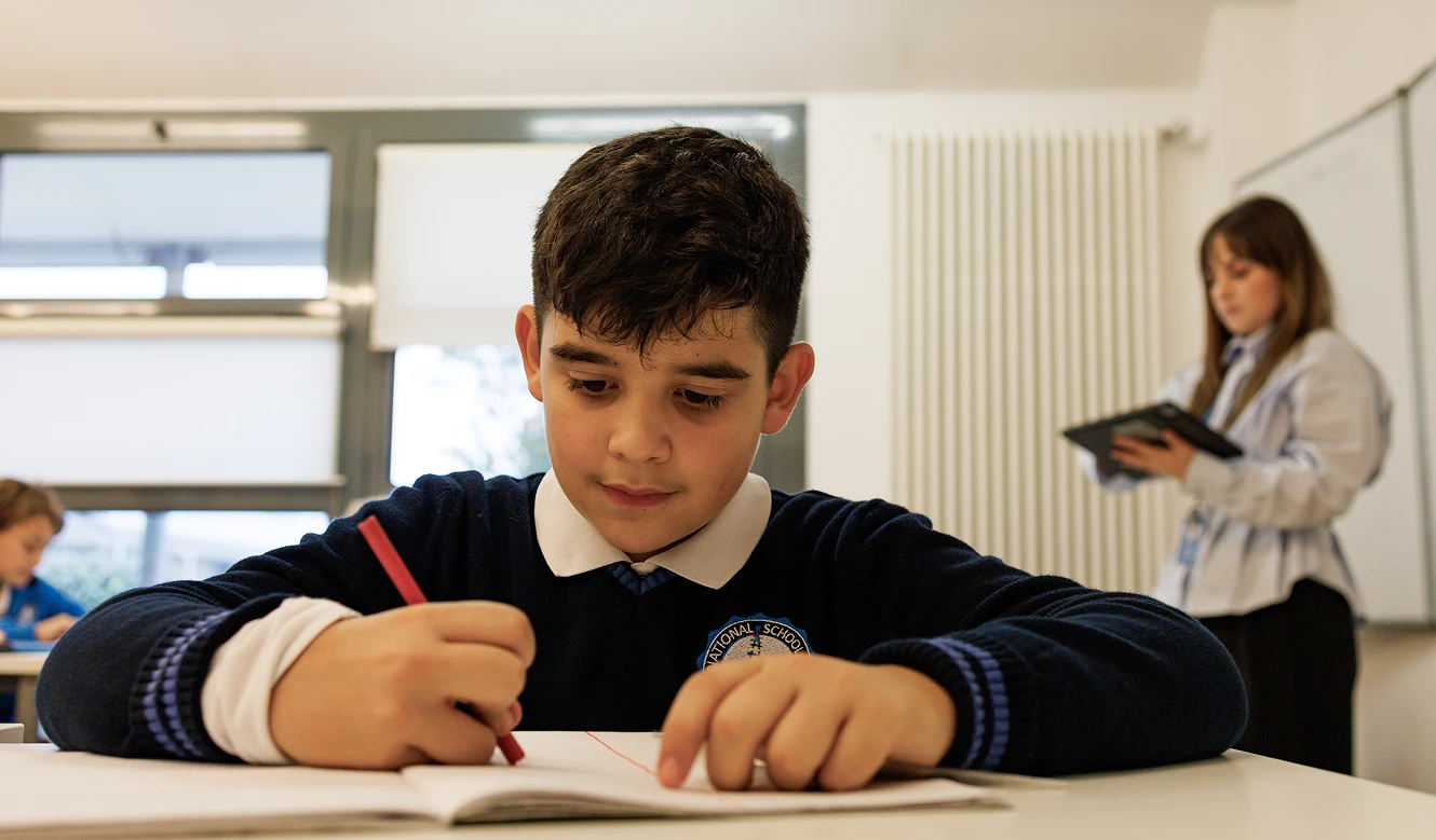 Foto a mezzo busto di un alunno di IST, istituto di primo piano nelle scuole paritarie a torino. Il ragazzo bruno con un pullover blu della scuola sta scrivendo con una penna rossa su un quaderno