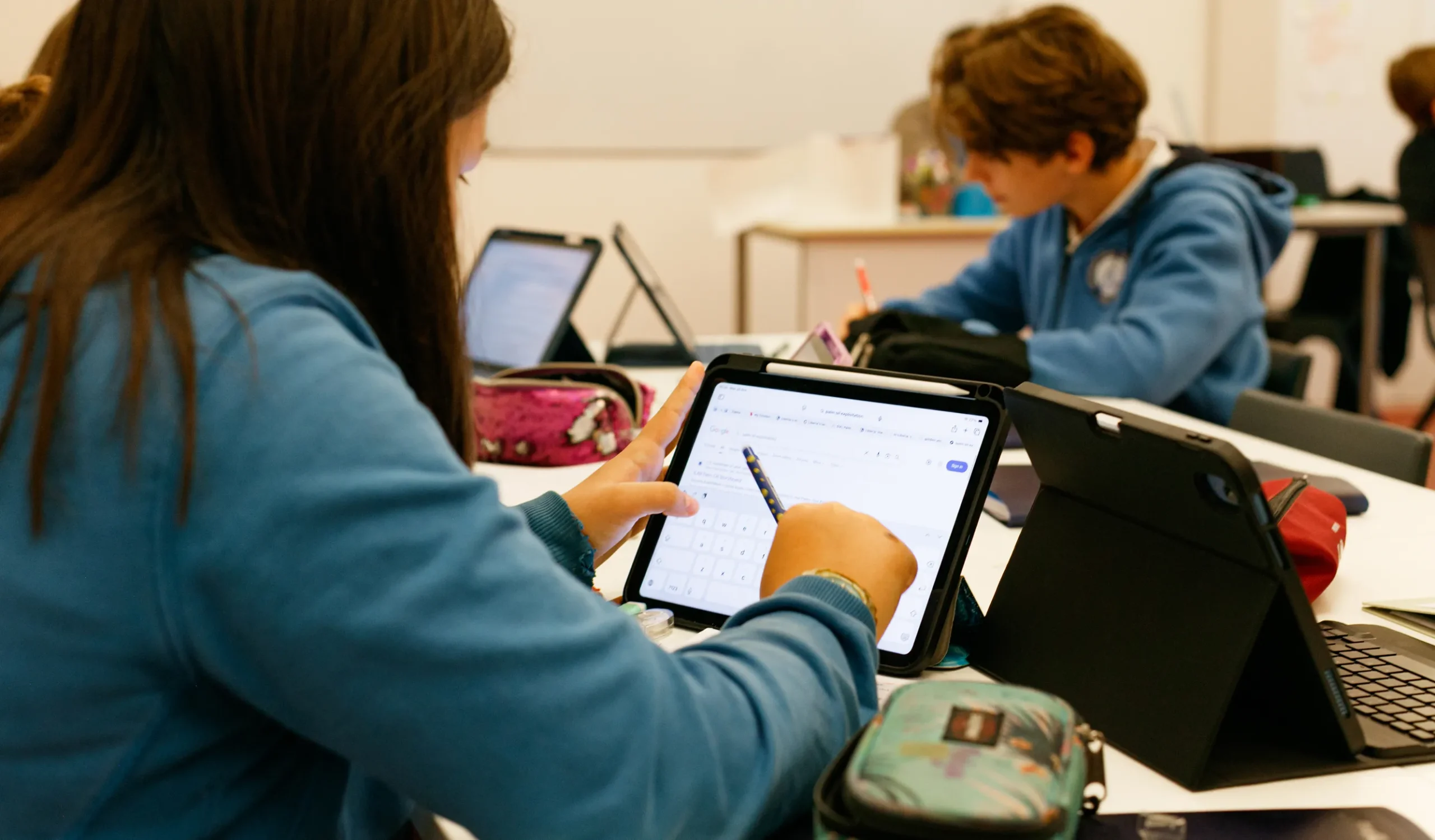 Una ragazza di spalle e un ragazzo di fronte a lei, con le felpe azzurre della divisa IST, lavorano con tablet in un’aula scolastica