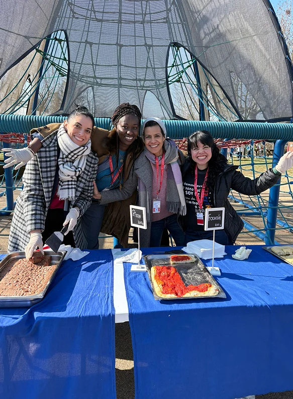 Quattro giovani donne sorridenti in abiti invernali offrono dolci e pizza da teglie posate su banchi con tovaglie azzurre durante un evento della parent teacher association