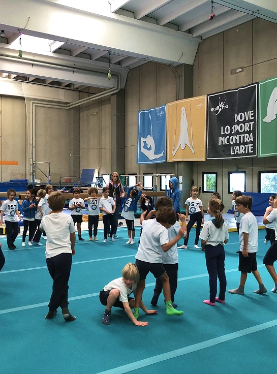Numerosi scolari con pantaloni blu navy e felpe bianche dell’IST svolgono attività sportive di gruppo in un’ampia palestra da atletica