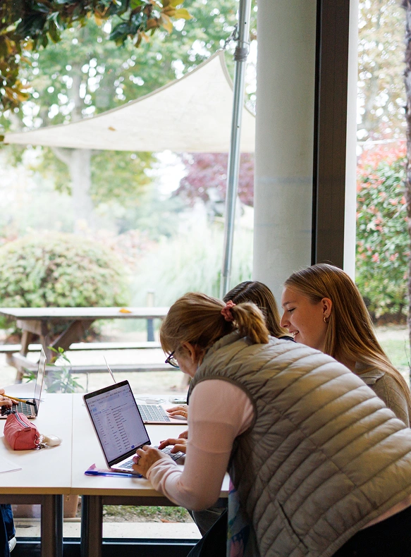 Un’insegnante dell’IST con un gilet beige mostra a due studentesse una pagina di dati da un laptop nel corso degli open day dell’università a torino. Attraverso le ampie finestre si vede un parco verde