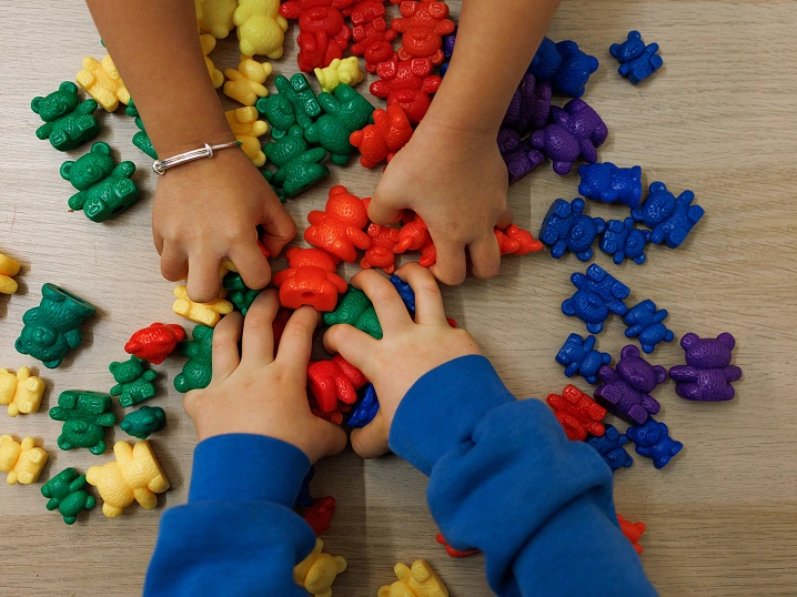 Le mani di due bambini giocano con orsetti di plastica colorati di rosso, giallo, verde, blu e viola. Lo scolaro in basso ha una felpa azzurra, l’altro un fine braccialetto metallico