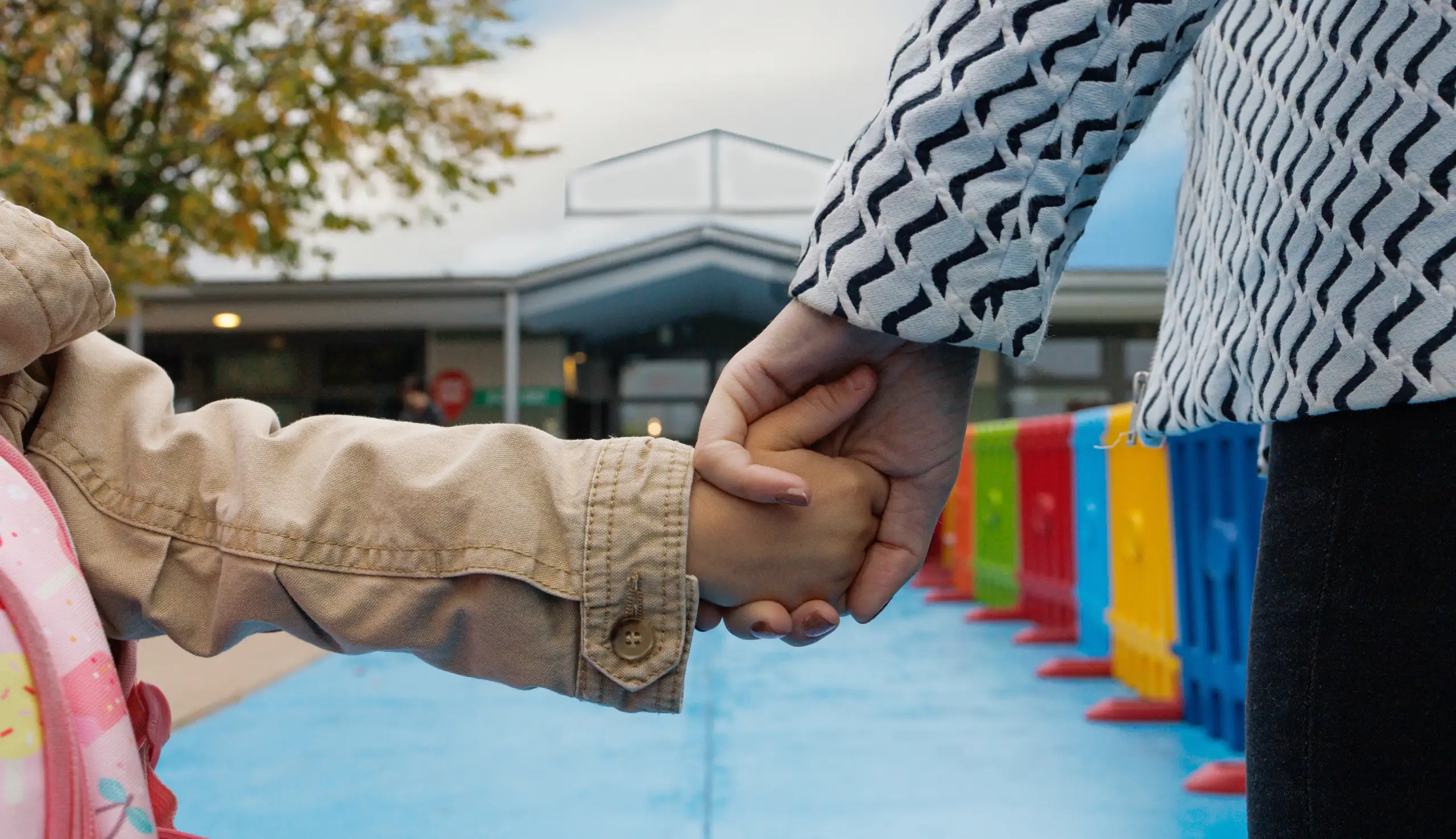 Una bambina di spalle, con una giacca beige e uno zainetto rosa, tiene la mano destra in quella di una donna vestita di bianco e nero che la accompagna verso l’ufficio ammissioni della scuola internazionale di IST